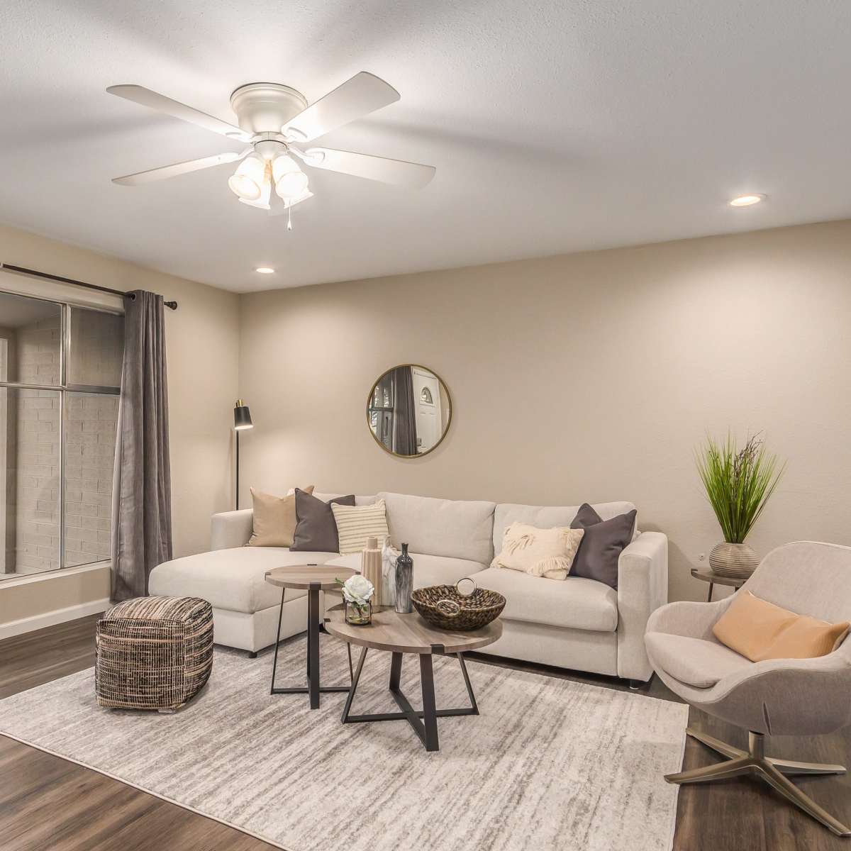 A living room features a beige sectional sofa, a round coffee table, and a large window with gray curtains.