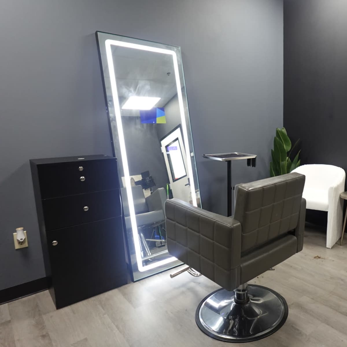 A minimalist salon interior featuring a gray chair, a large illuminated mirror, a small side table, and a storage cabinet against a dark wall.