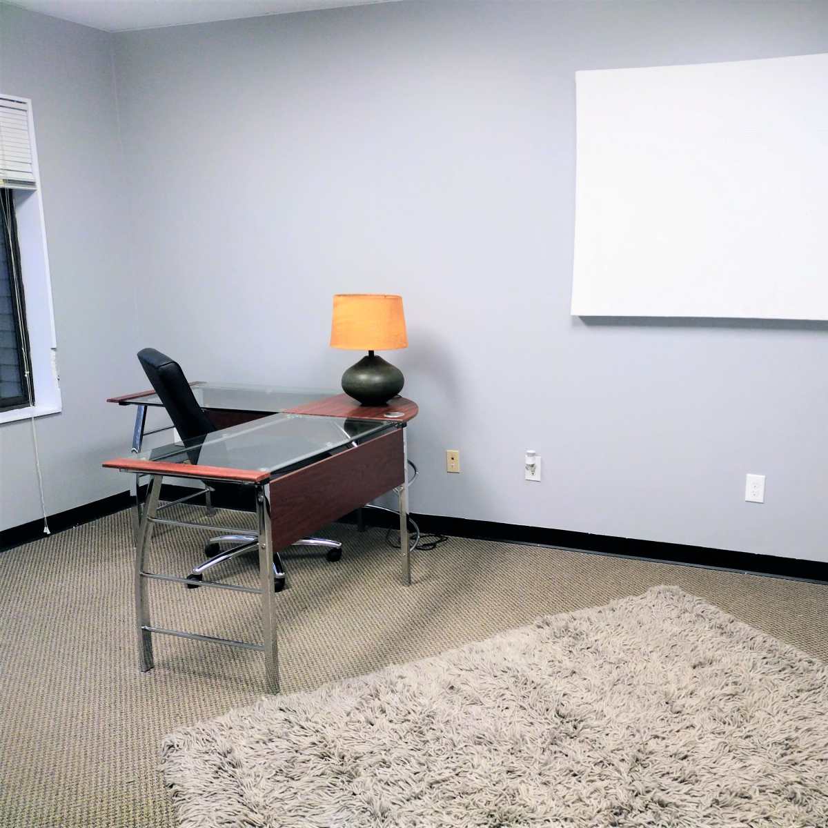 A sparsely furnished office with a glass desk, a black chair, an orange lamp, and a large blank canvas on the wall.