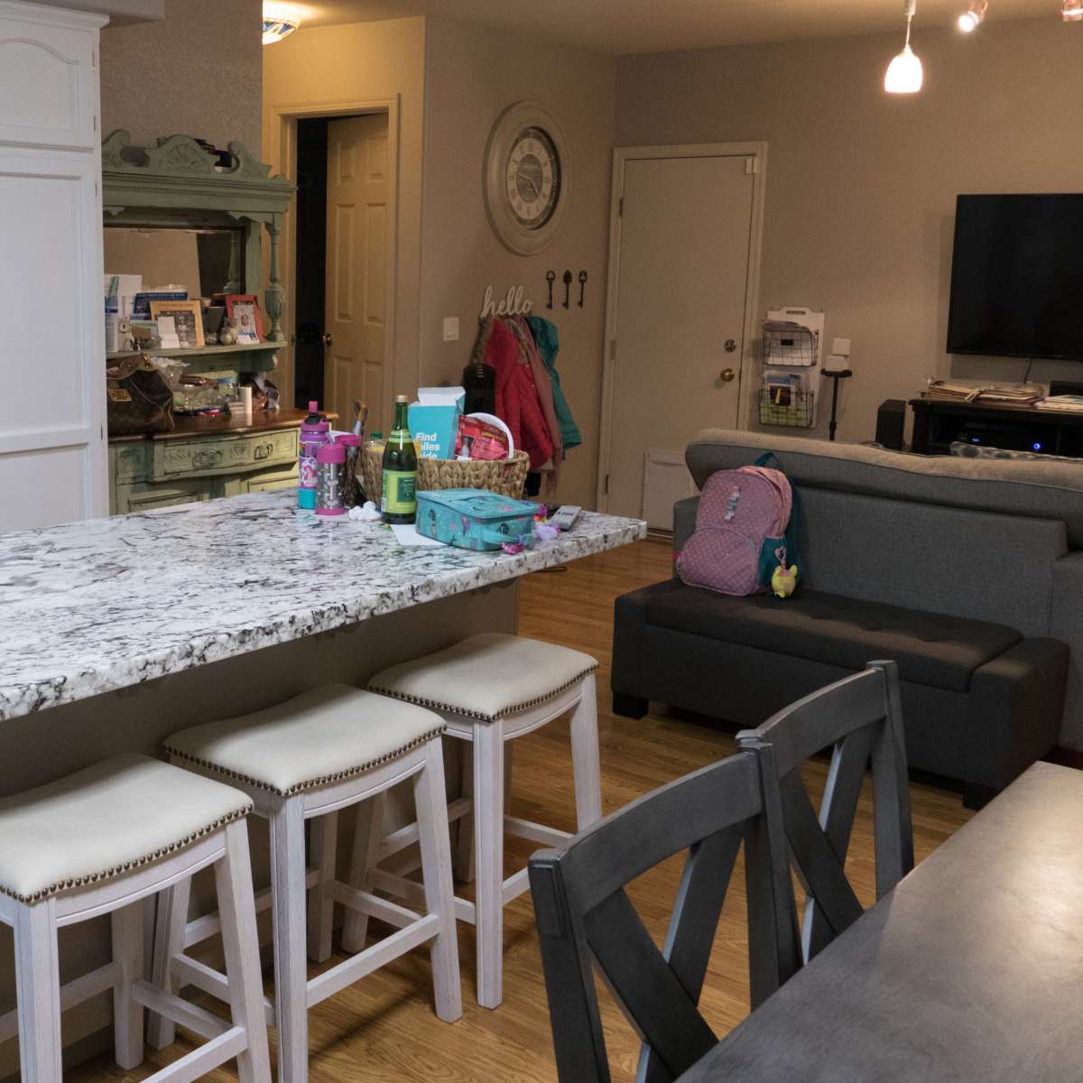 A kitchen and dining area with a granite-topped island, three white stools, and various items on the countertop, along with a couch and television in the background.