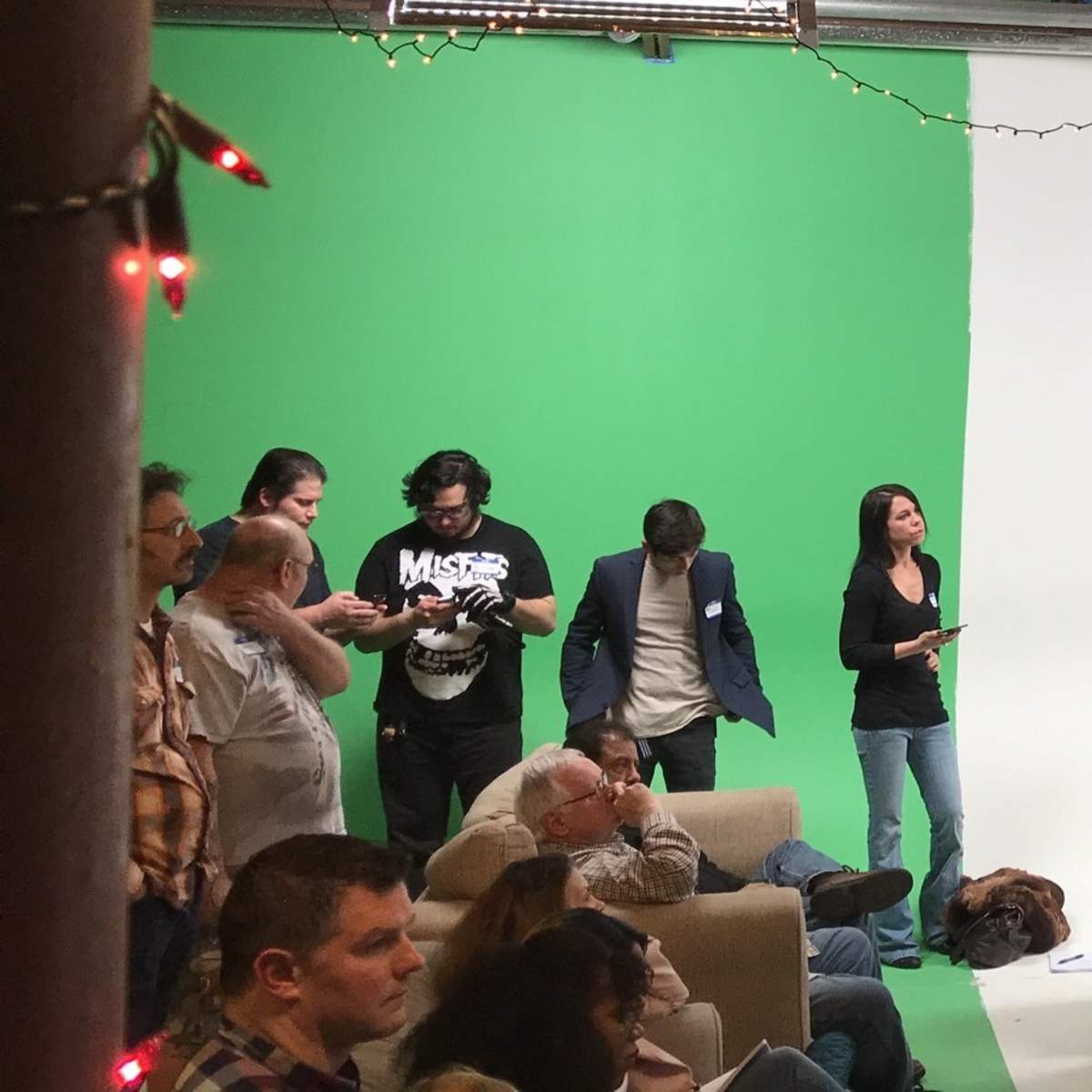 A speaker stands on a table in front of a green screen while an audience listens and takes notes.