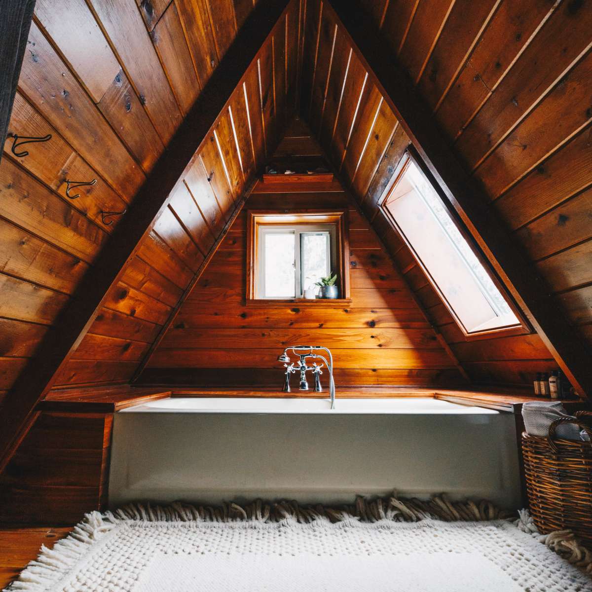 A triangular wooden bathroom with a freestanding bathtub and a window above it, set against a textured rug on the floor.