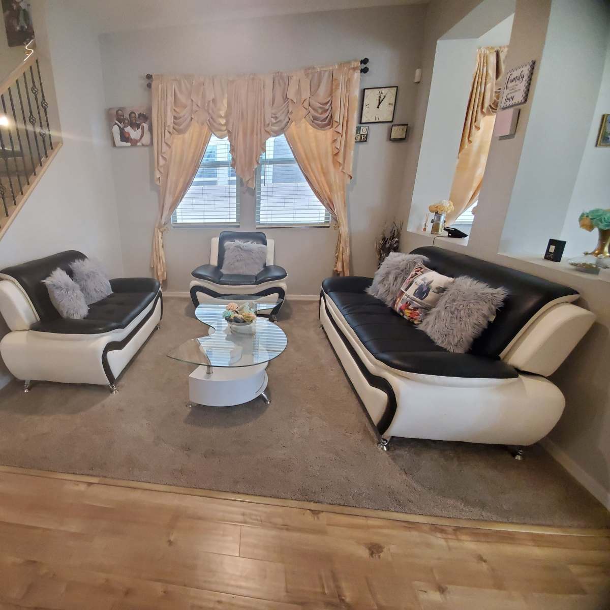 A living room with two modern black and white sofas, a glass coffee table, and large windows with drapes, adjacent to a staircase with a wooden railing.