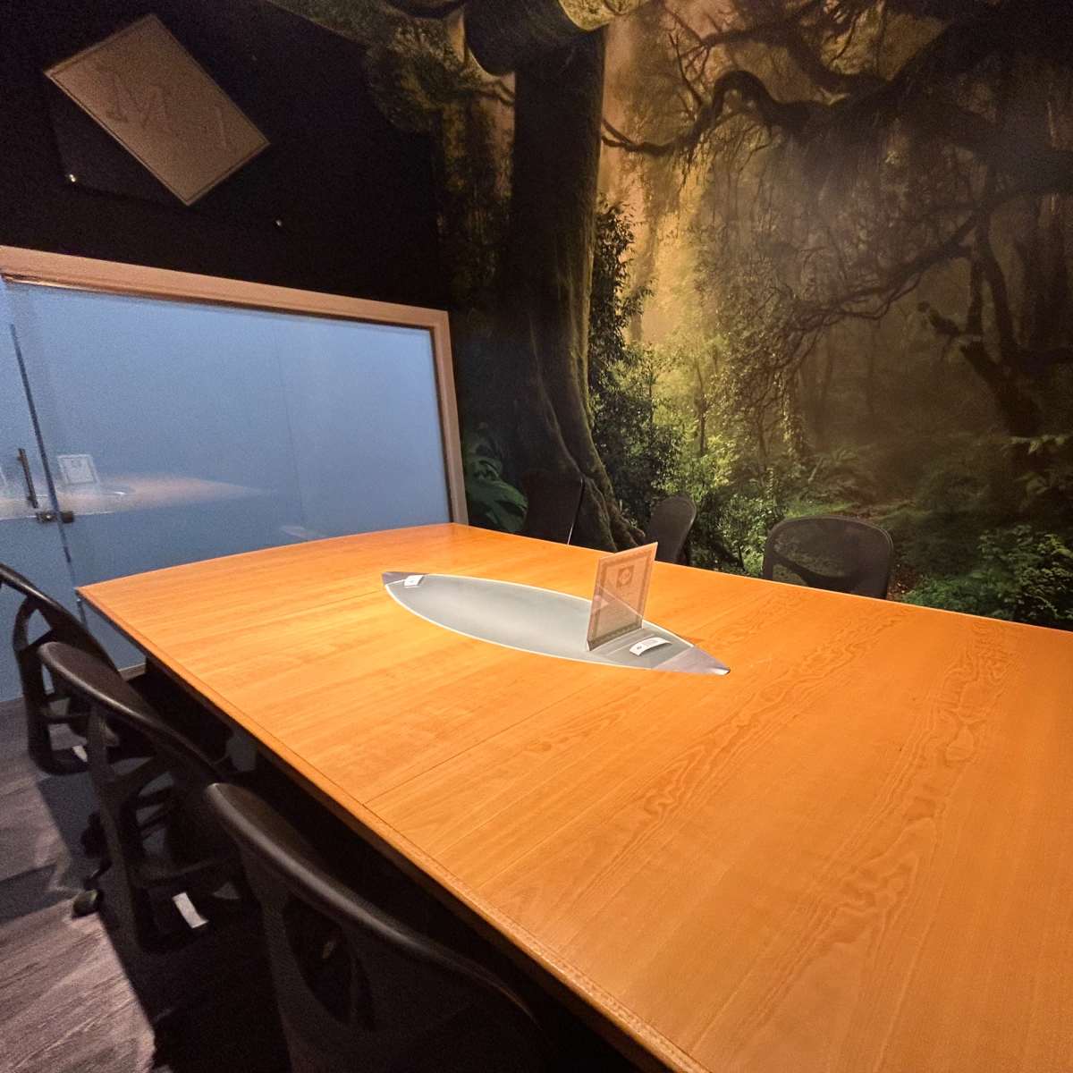A conference room features a large wooden table surrounded by black chairs, set against a mural of a forest landscape.