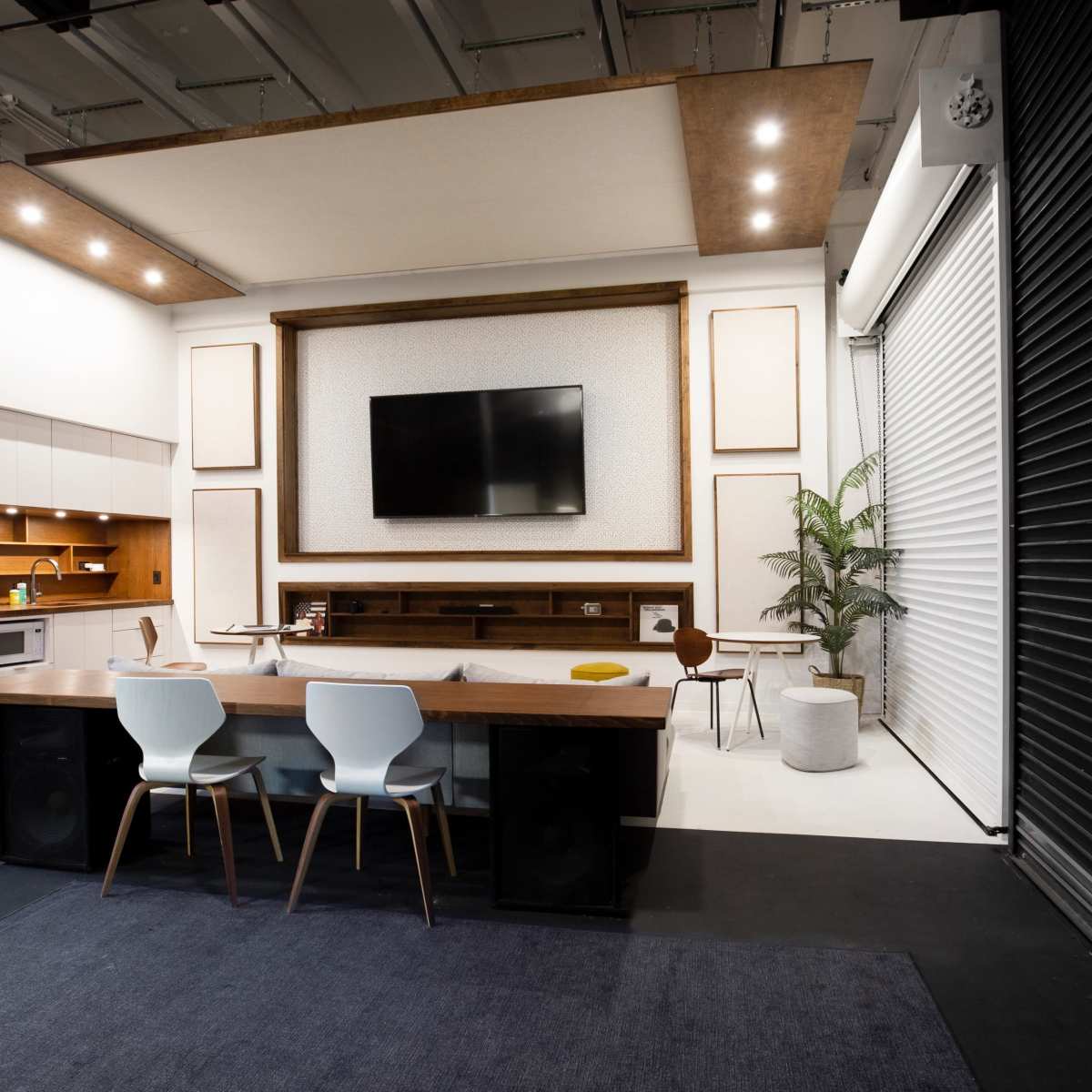 A modern indoor space featuring a kitchen area, a large television mounted on a wall, a dining table with chairs, and a potted plant.