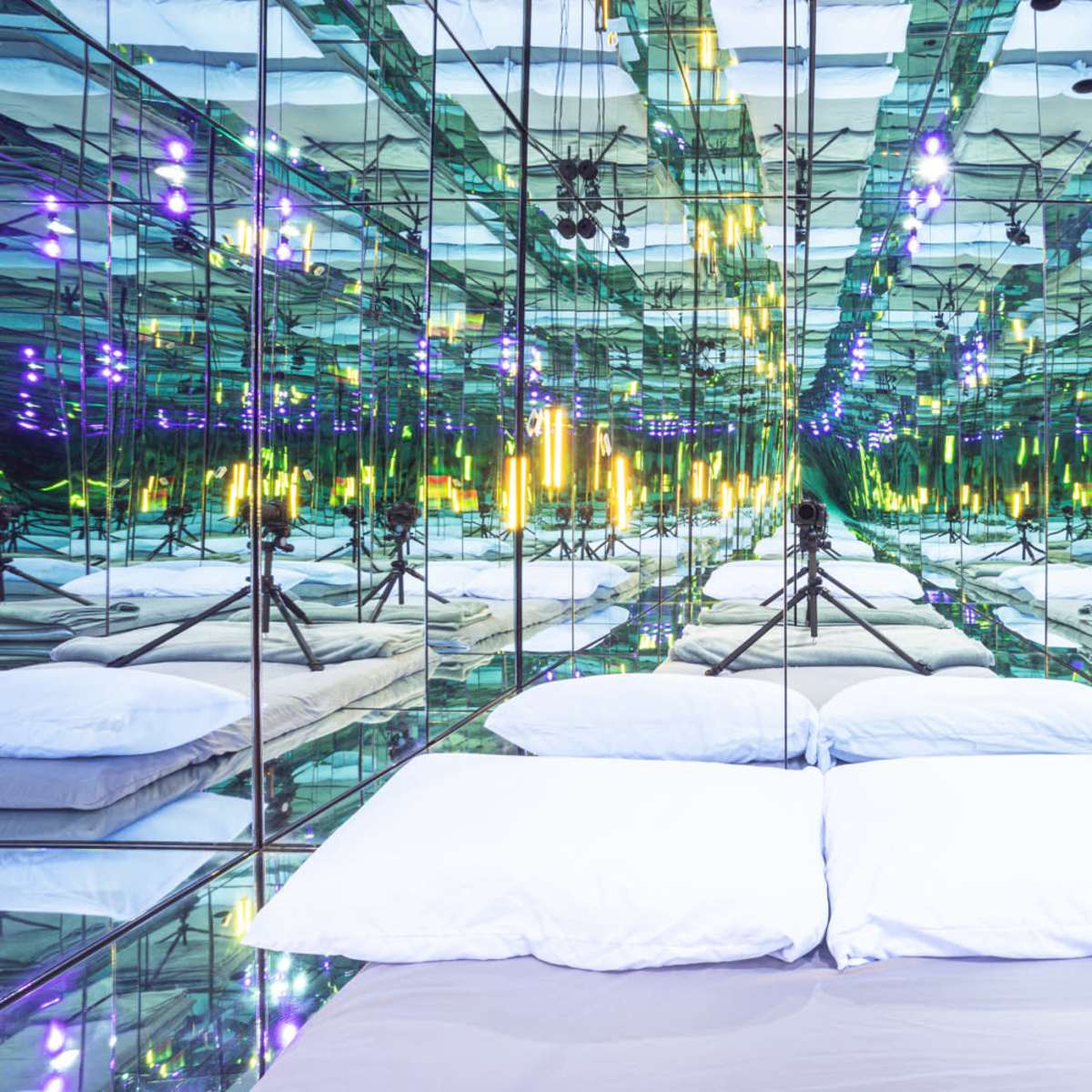 A mirrored room with a bedding area featuring two white pillows and multiple reflections of colorful lights.