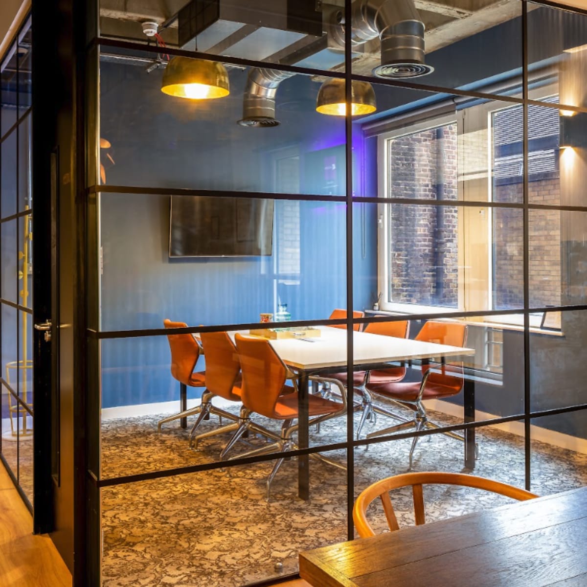 A modern conference room with a glass wall, featuring a table surrounded by leather chairs and wooden flooring.