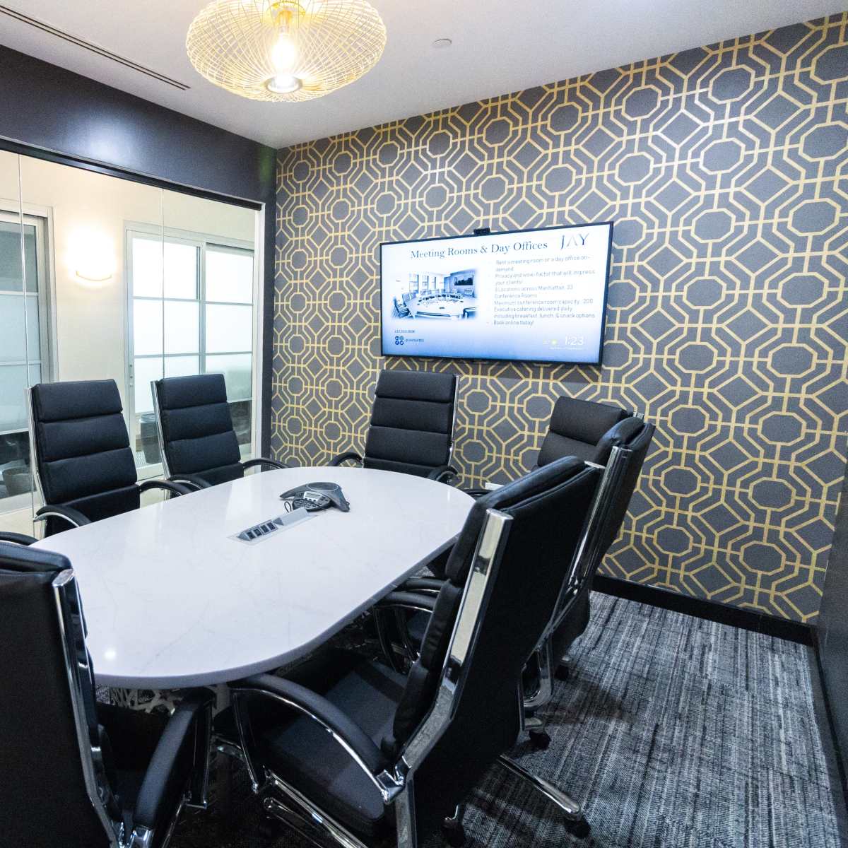 A modern conference room features a large oval table surrounded by black leather chairs, with a wall-mounted screen displaying information and a patterned wallpaper.