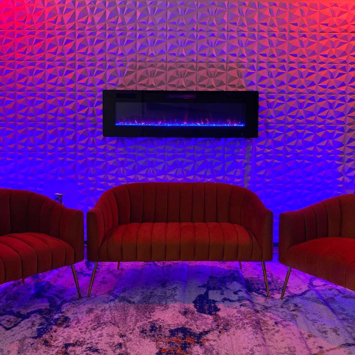 The scene features three red velvet chairs arranged near a modern wall-mounted electric fireplace, illuminated with blue and red lights against a textured wall.