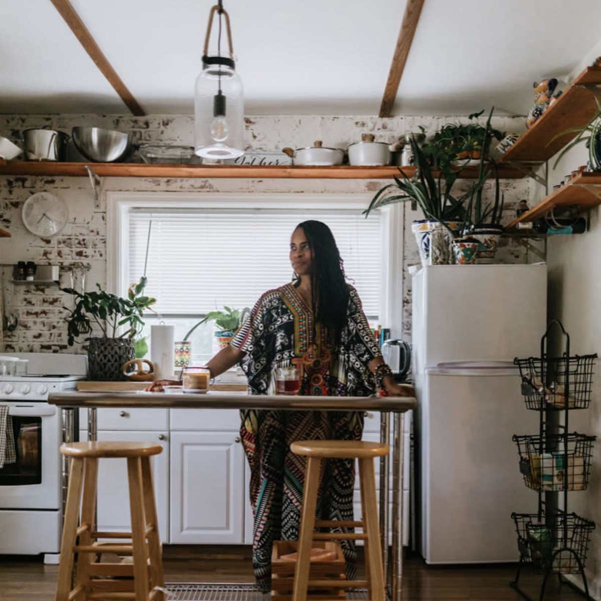 A woman stands at a kitchen island in a rustic kitchen filled with plants and wooden shelves.