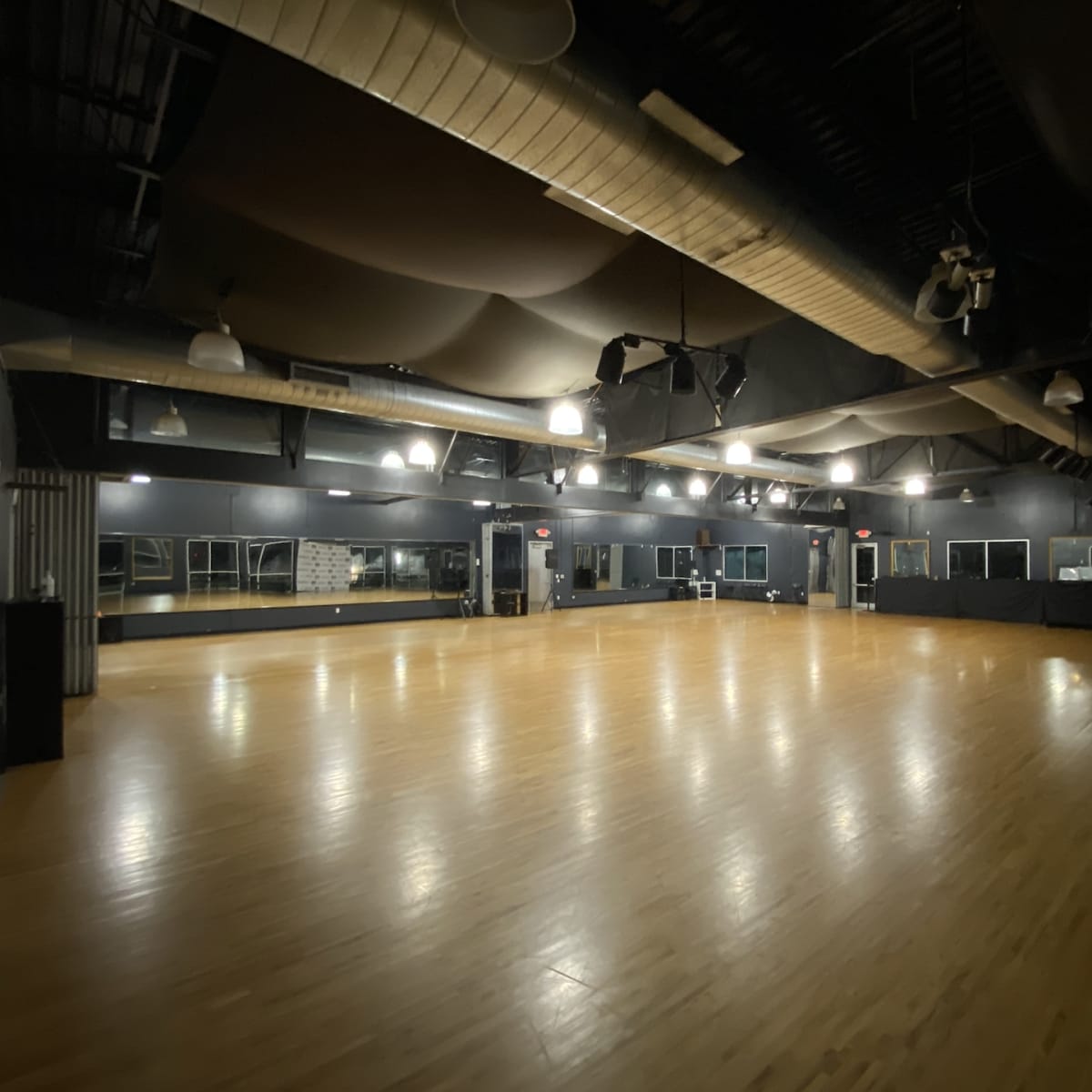 A spacious dance studio with polished wooden floors, dim lighting, and large mirrors along one wall.