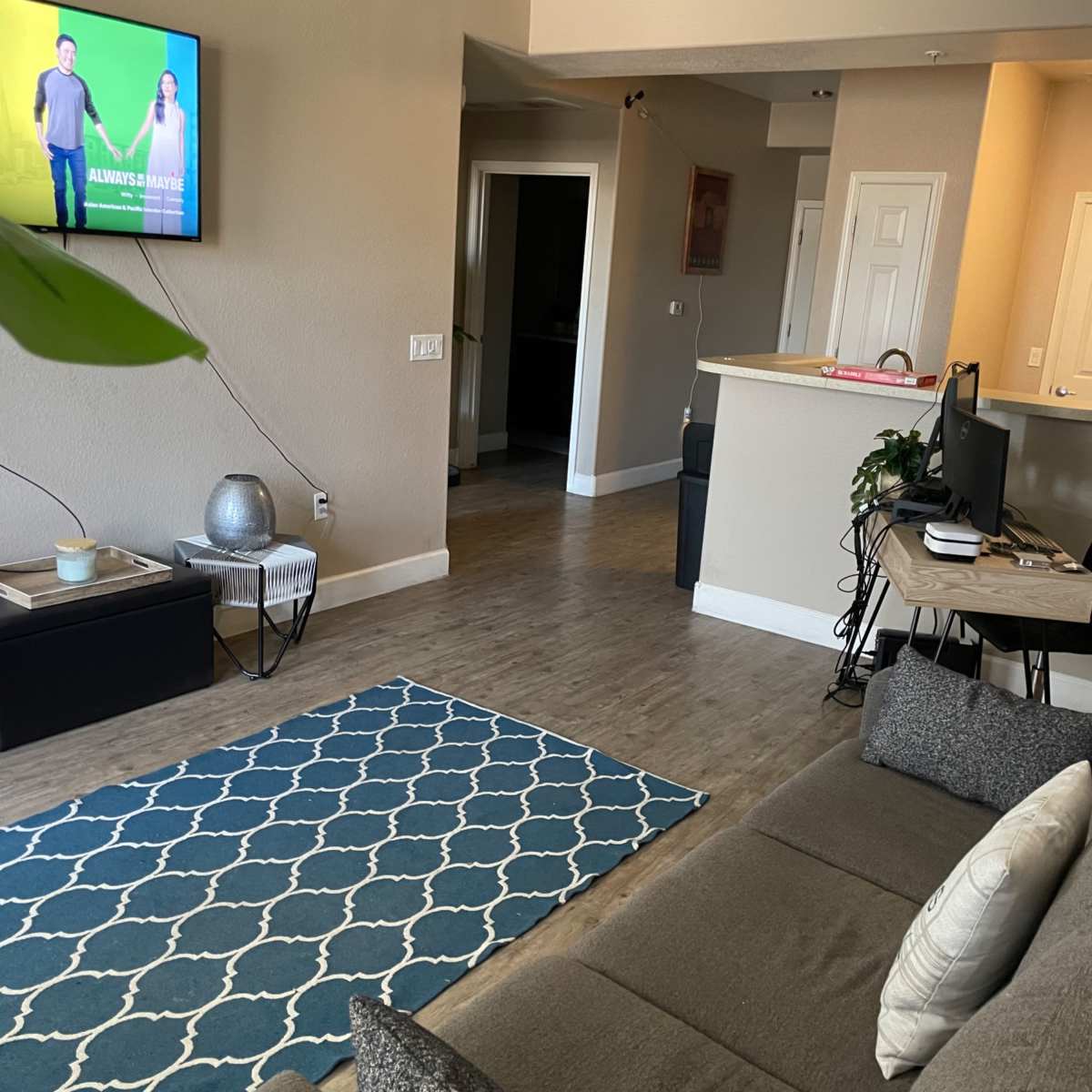 A living room with a sofa, a patterned rug, a television mounted on the wall, and a small desk setup in the corner.