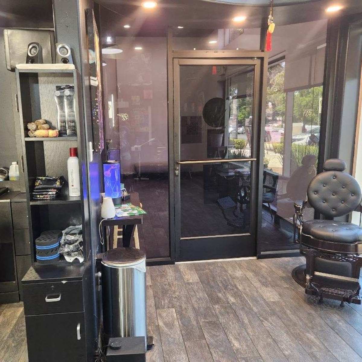 A sleek barbershop interior featuring two black barber chairs, a retail shelving unit with grooming products, and a glass entrance door.