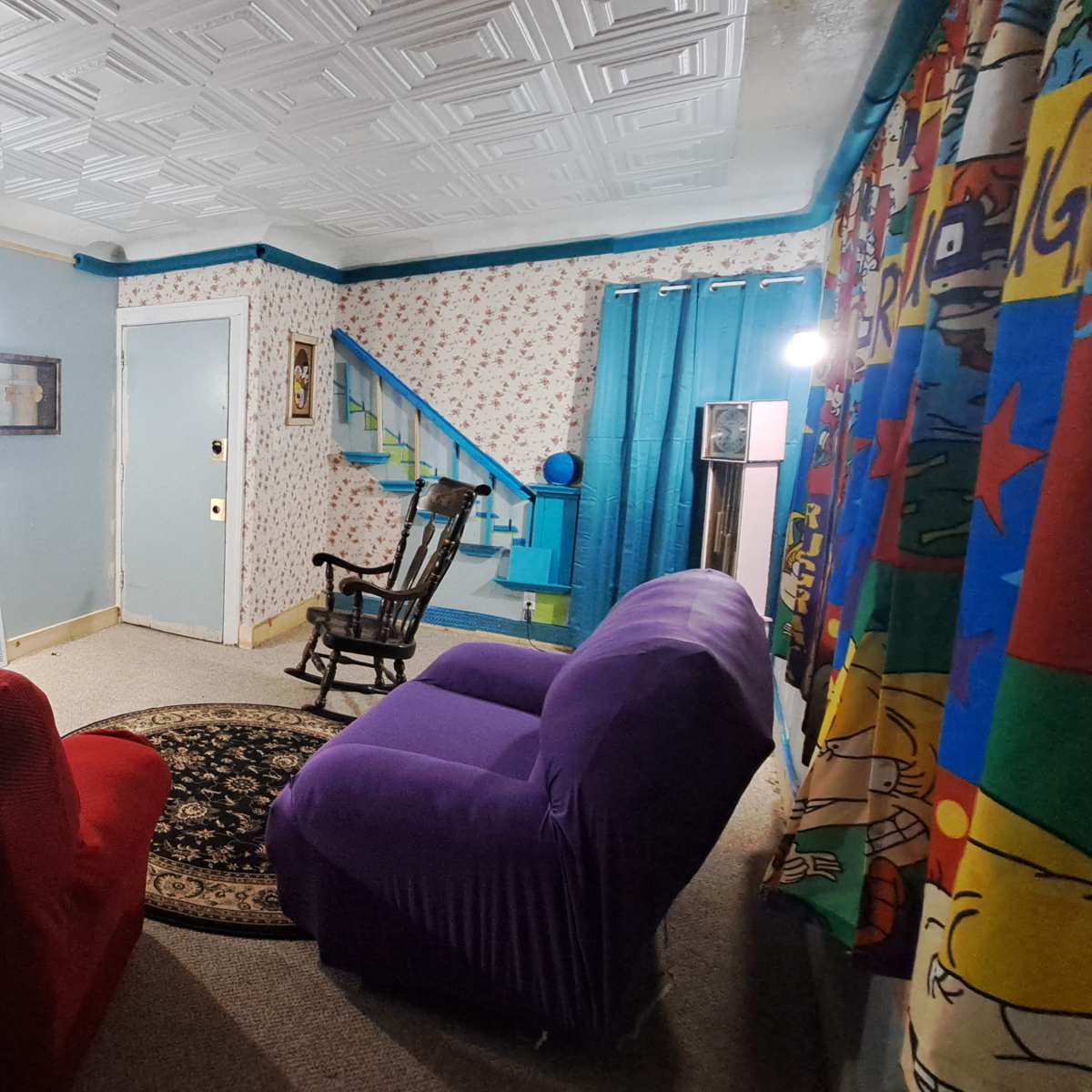 The living room features two colorful armchairs facing a patterned area rug, with blue curtains and floral wallpaper.