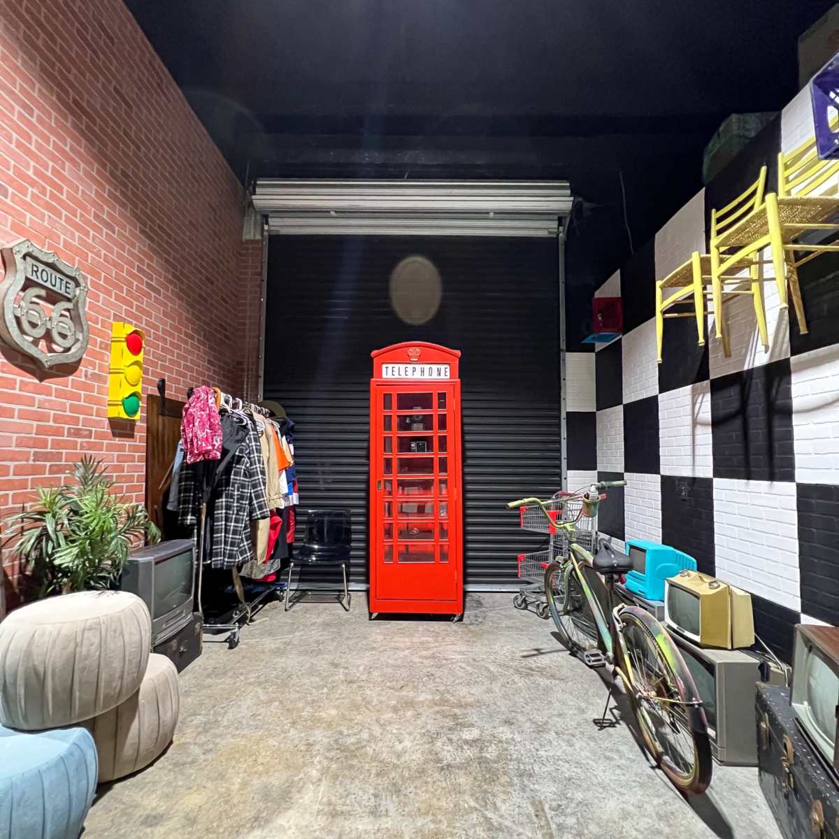 A vintage red telephone booth in a garage filled with various clothing items, bicycles, and furniture against a backdrop of brick and checkered walls.
