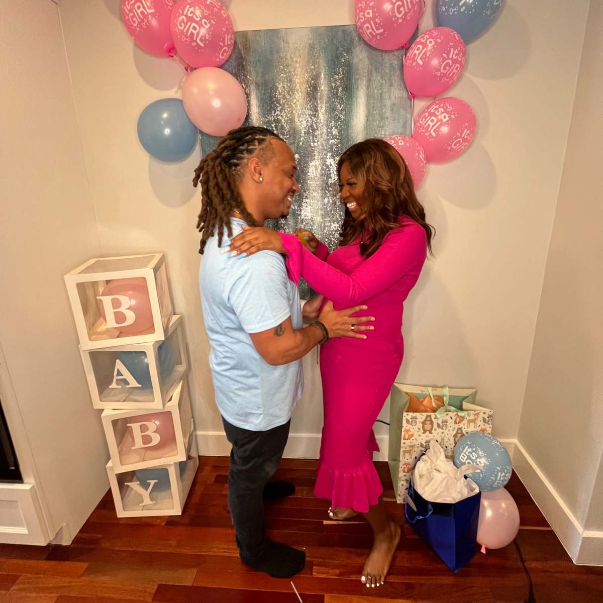 A couple stands together in a decorated room with pink and blue balloons and a display that reads "BABY."