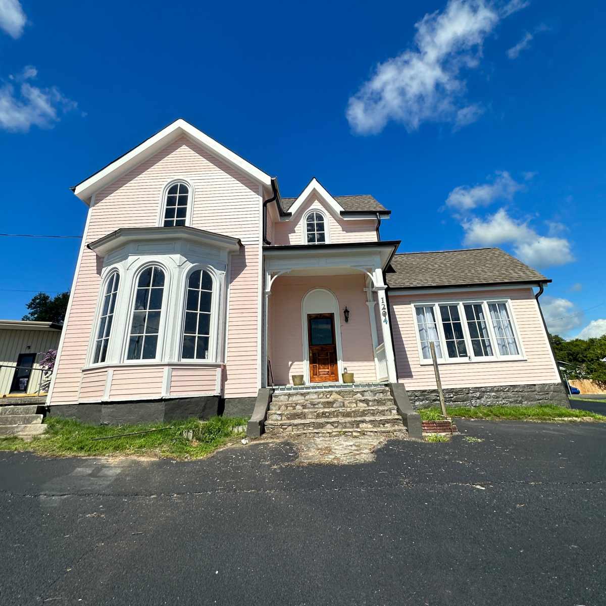 A light pink house with large arched windows and a gray shingled roof stands against a blue sky with scattered clouds.