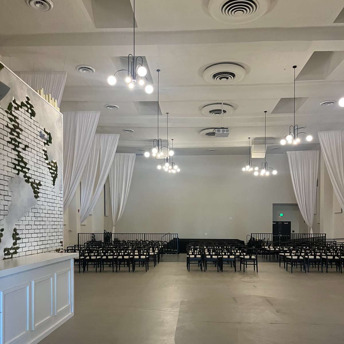 A spacious event venue with white drapes, pendant lights, and rows of black chairs arranged in front of a blank wall.