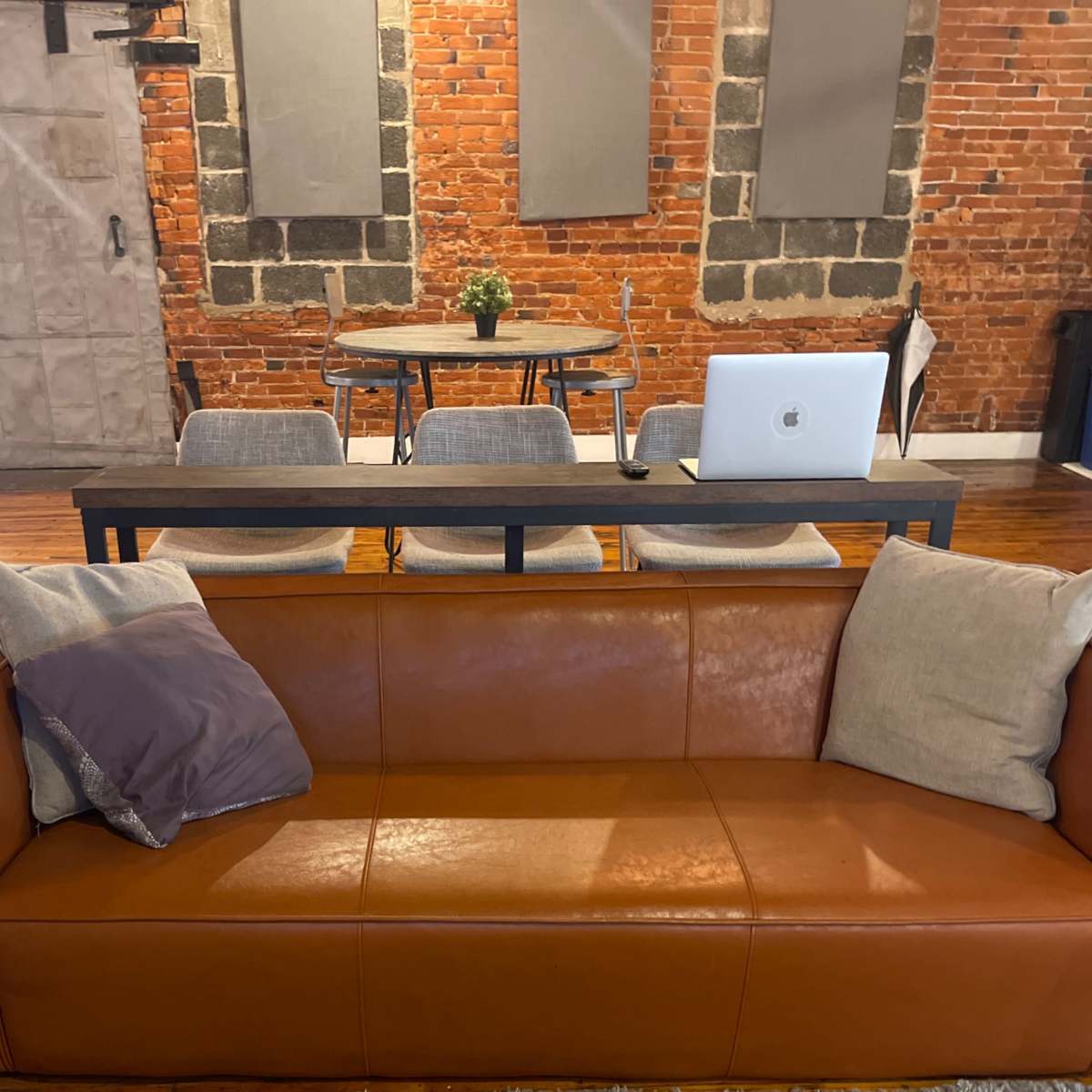 A brown leather couch with gray cushions in a room that has exposed brick walls and a wooden table with a laptop on it.