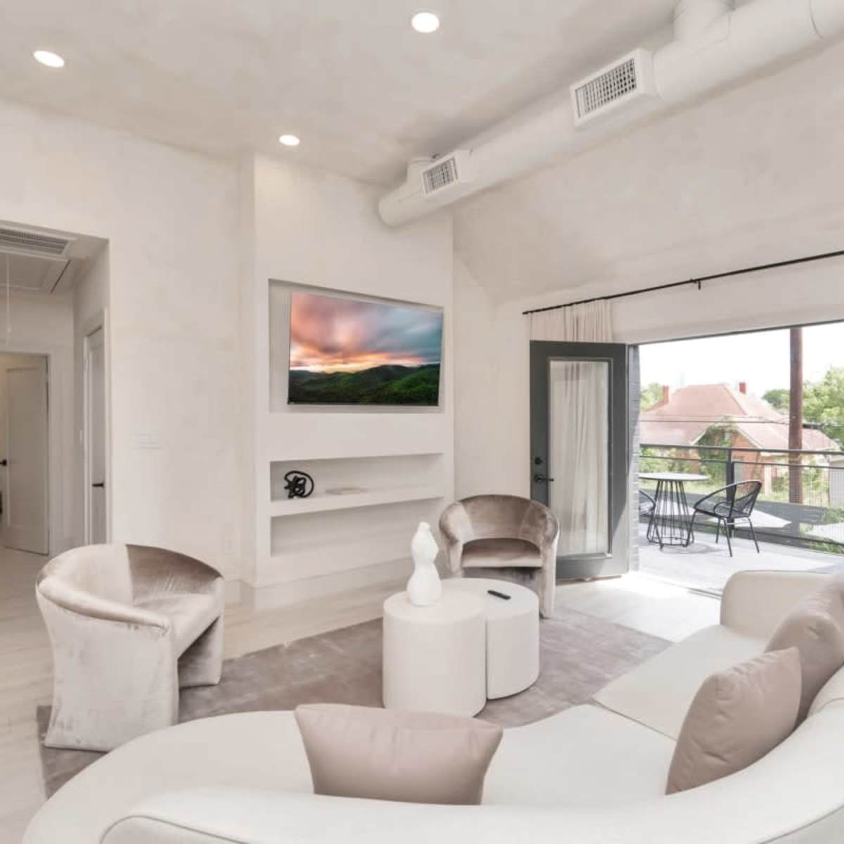 A modern living room features a curved sofa, two armchairs, a minimalist coffee table, and a wall-mounted TV, with large sliding doors leading to a balcony.