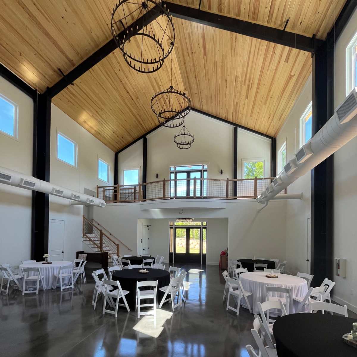 A spacious event hall with high ceilings, wooden beams, several round and rectangular tables, and a staircase leading to an upper level.