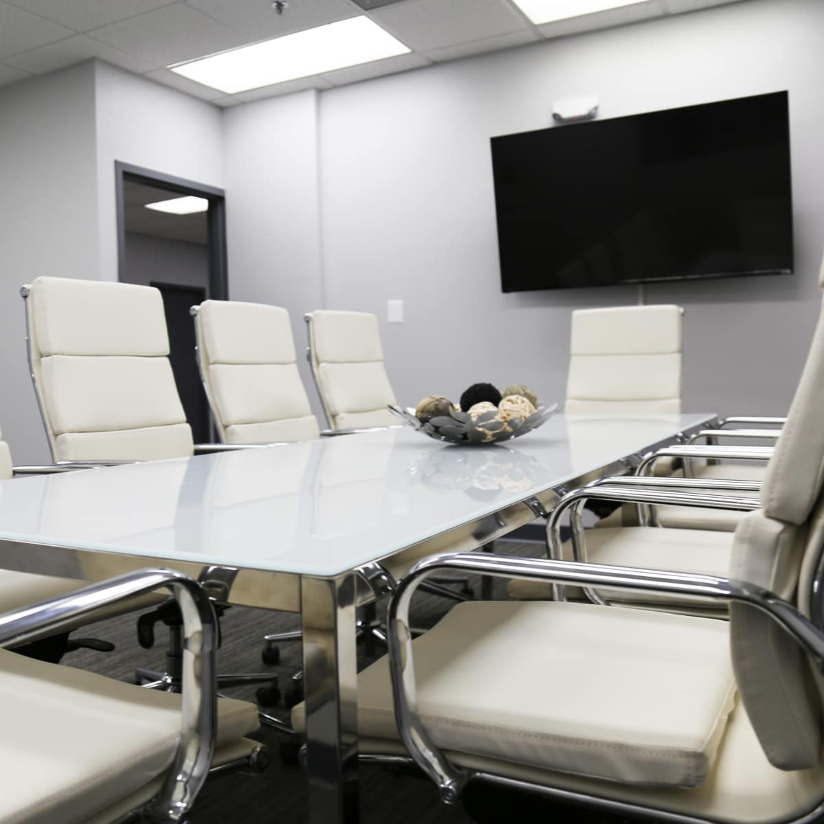 A conference room features a glass table surrounded by six white leather chairs and a wall-mounted television.