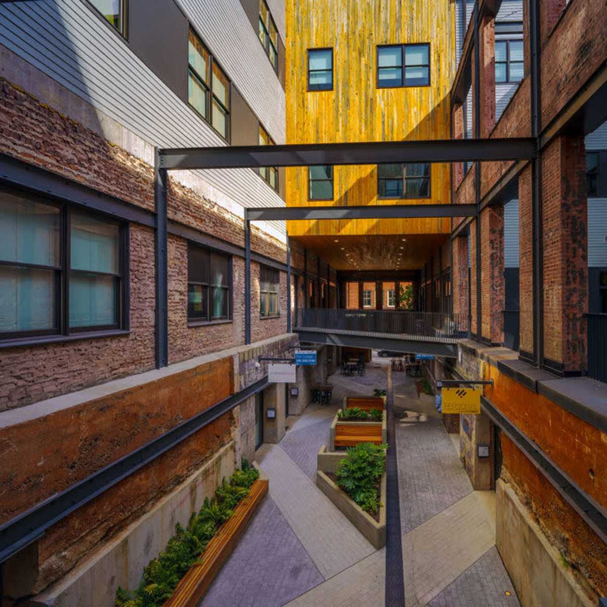 A modern urban courtyard featuring brick walls, wooden accents, and a pathway lined with greenery.