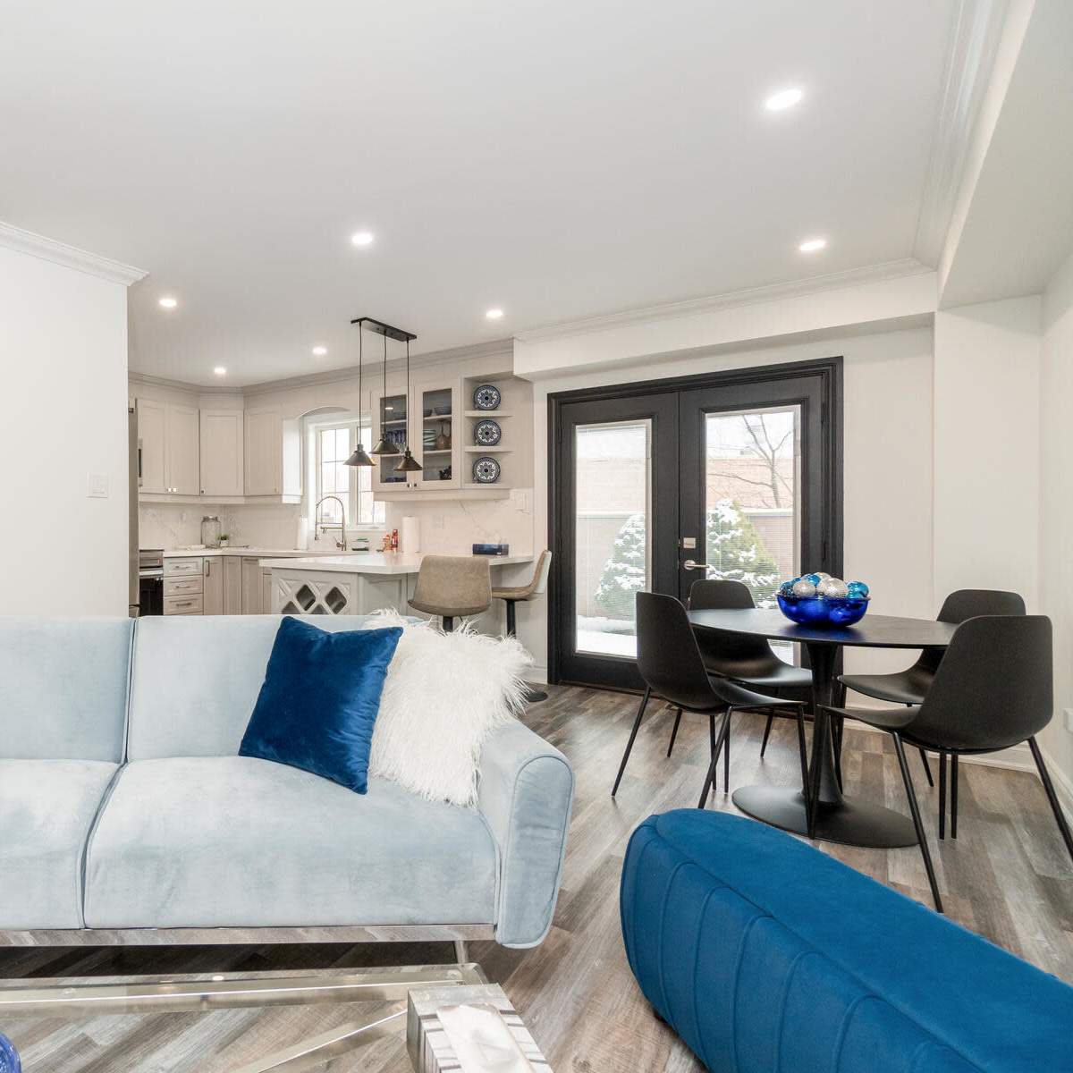 A modern living space features a light blue sofa with decorative pillows, a dining area with black chairs, and a kitchen visible in the background.