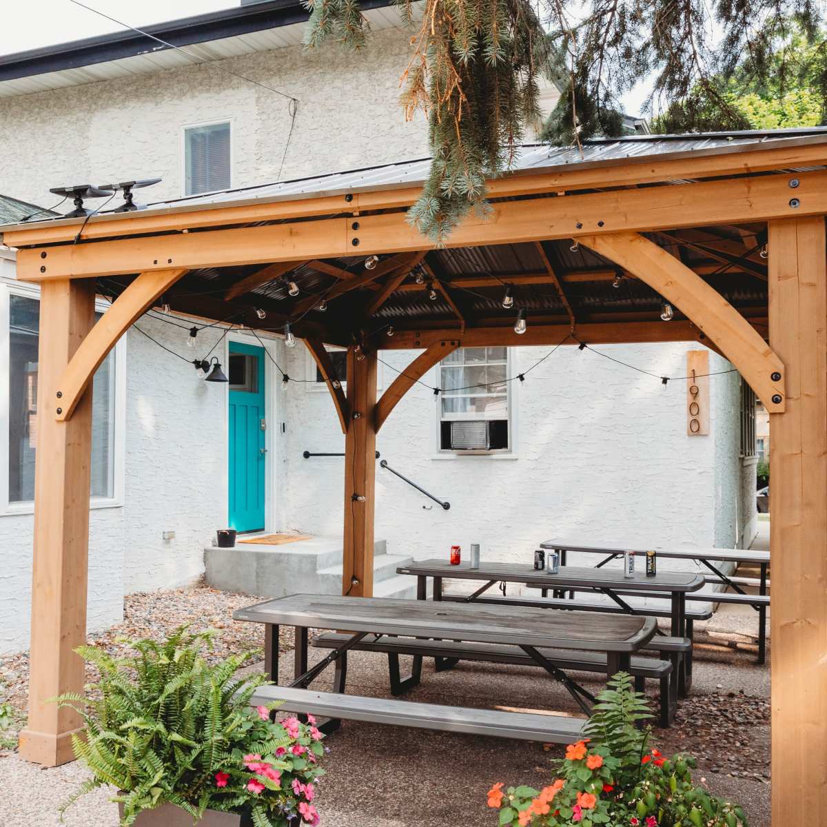 A wooden pergola with string lights provides shade over picnic tables in a gravel patio area beside a white house.