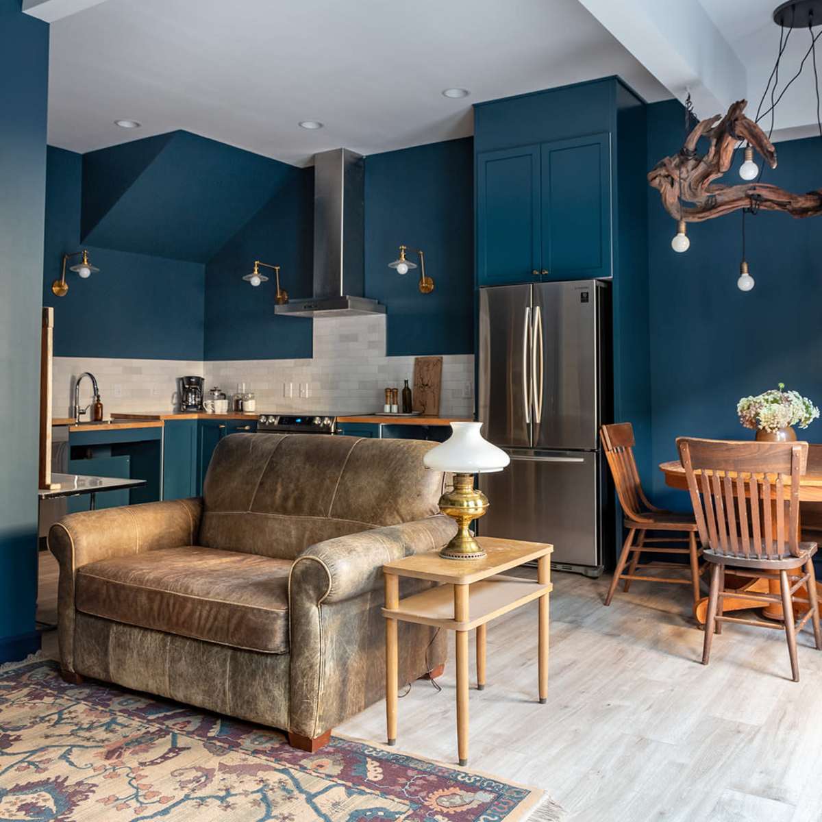 A modern kitchen and living area featuring a brown leather sofa, a wooden dining table with chairs, and blue cabinetry.