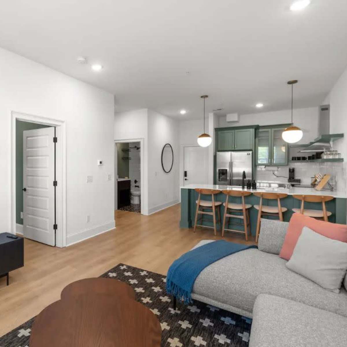 A modern living room and kitchen area featuring a flat-screen TV, a brown armchair, a gray sofa, and a kitchen with green cabinets and bar seating.