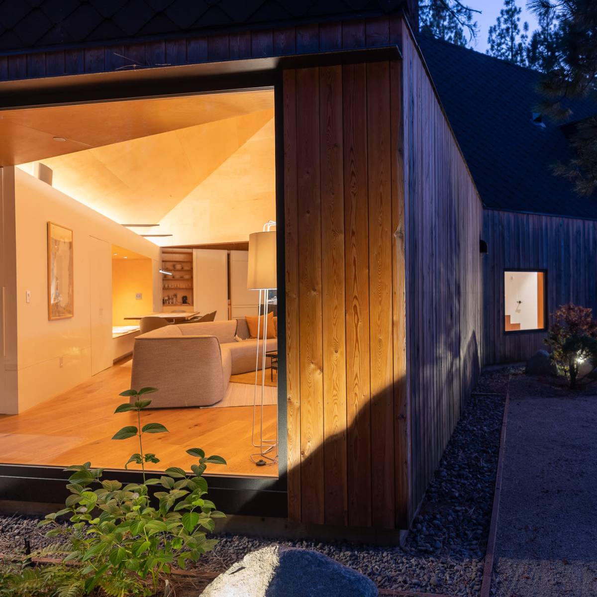 A modern house at dusk features a wooden exterior, large windows, and illuminated interior spaces that showcase a cozy living area.