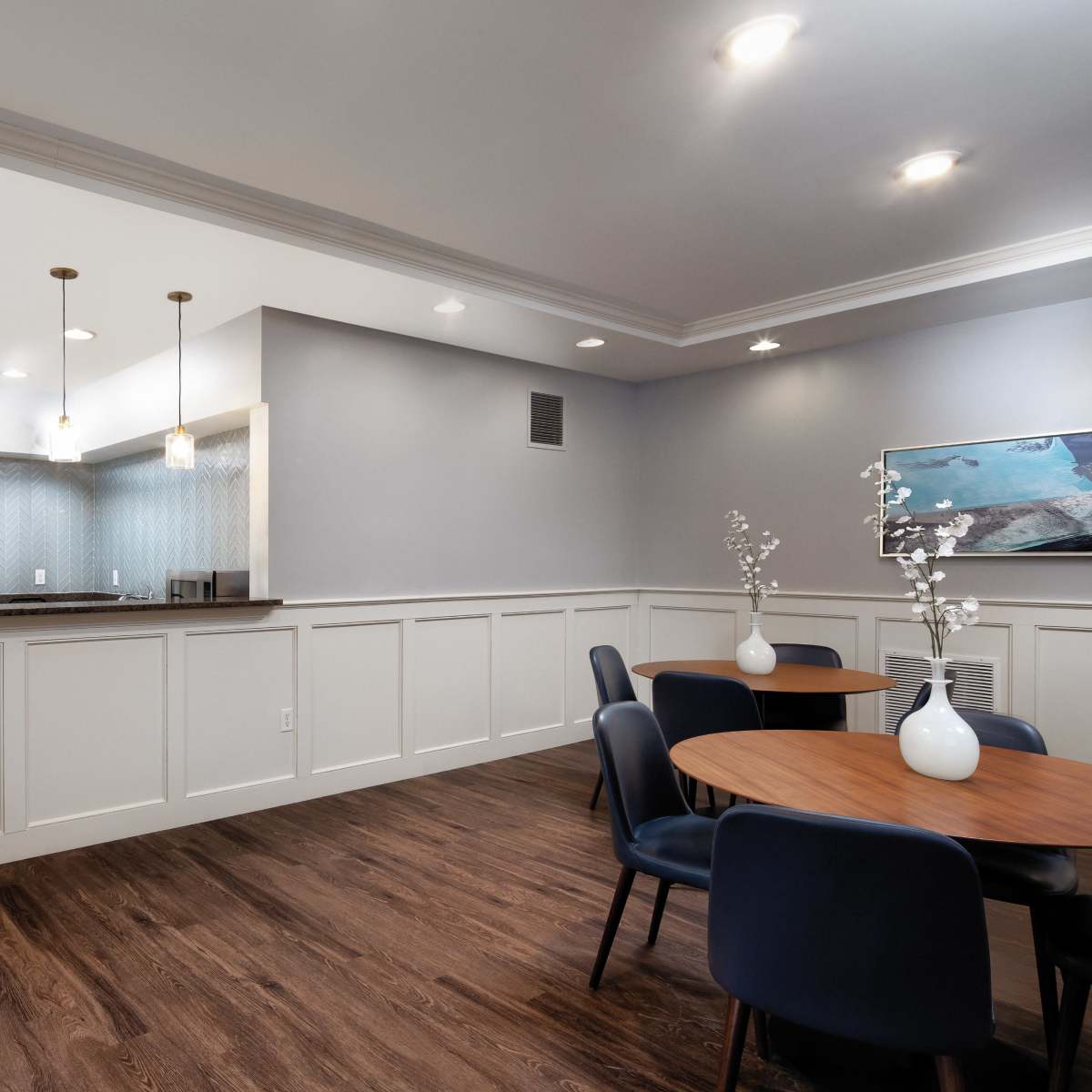 A modern kitchen and dining area with a light gray wall, wooden flooring, stainless steel appliances, and round tables with chairs.