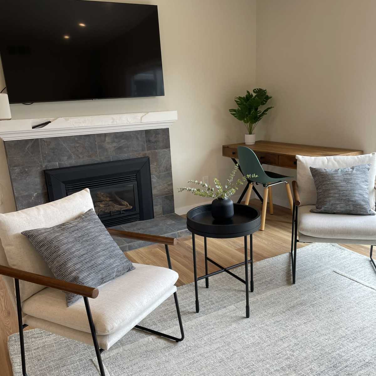 A living space with two upholstered chairs, a round black table, a fireplace, a television mounted on the wall, and a small dining table with a green chair.