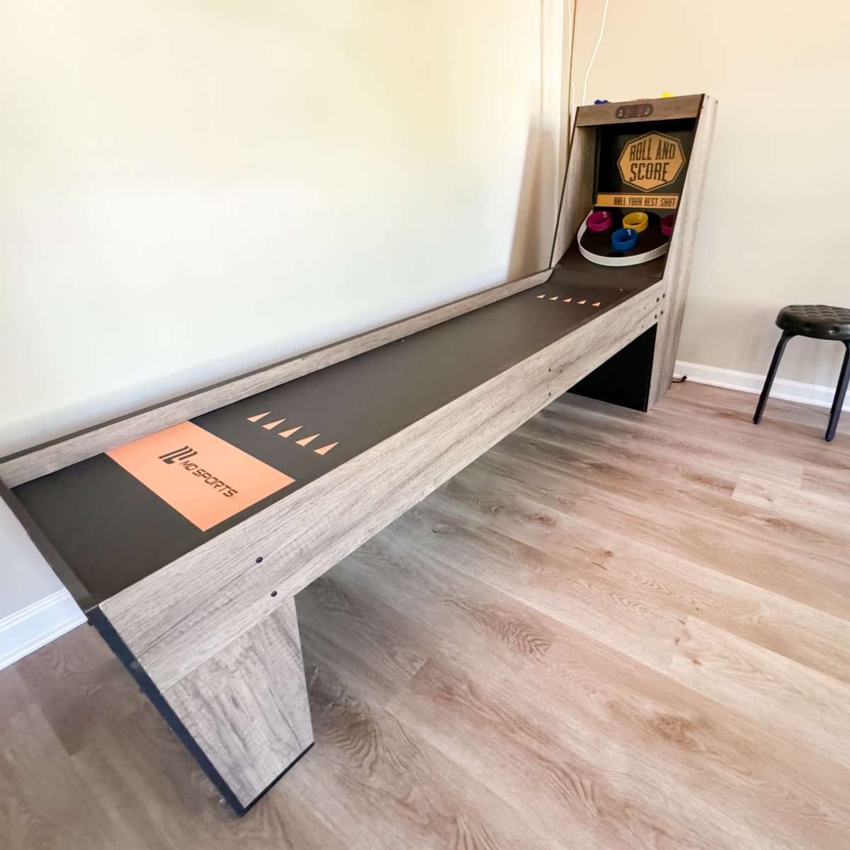 A wooden skee-ball arcade machine positioned against a wall with two black stools nearby.