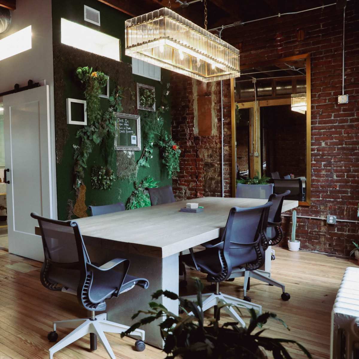 A modern office space featuring a large wooden table surrounded by black chairs, with exposed brick walls and greenery decor.