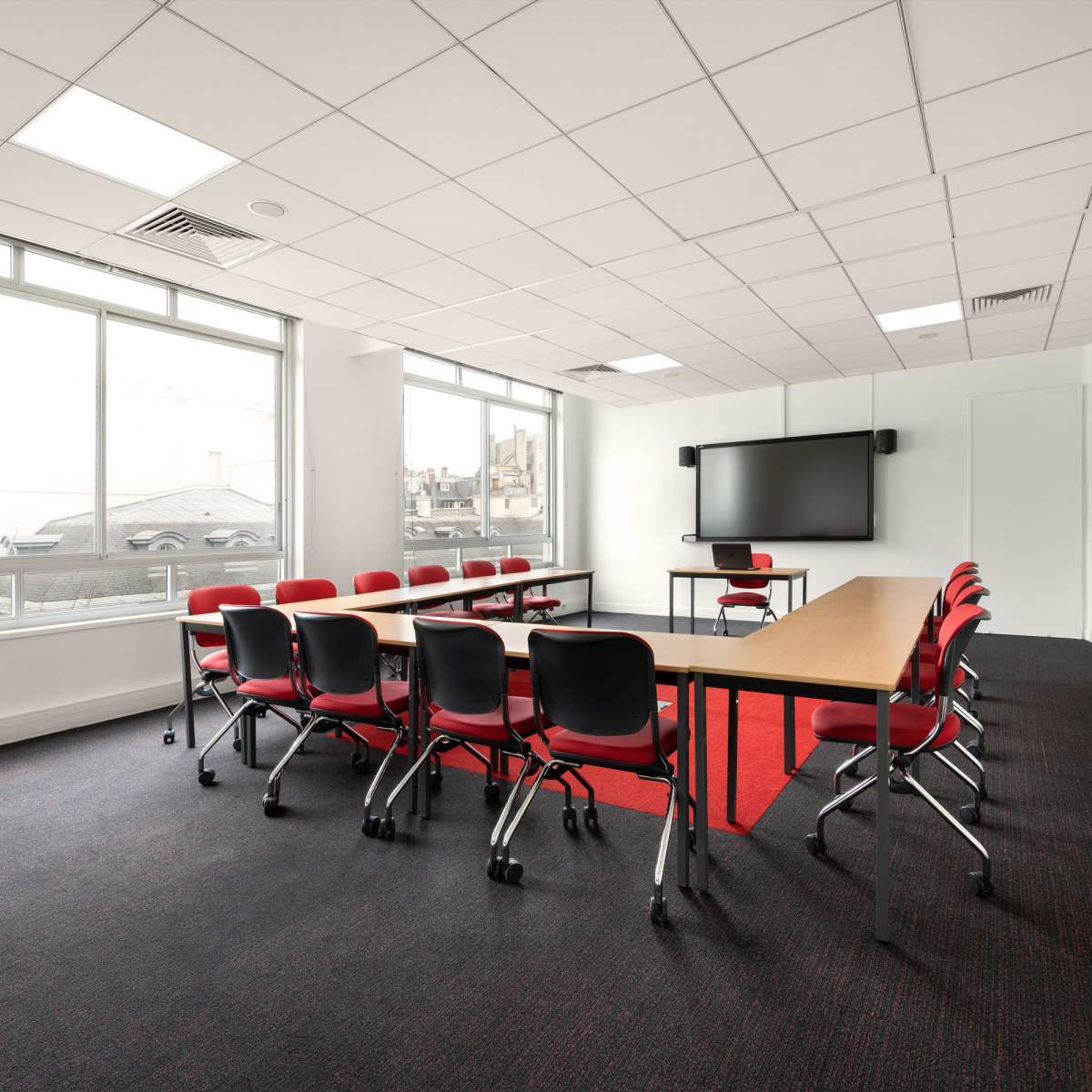 A conference room with a long table surrounded by red chairs, large windows, and a wall-mounted screen.