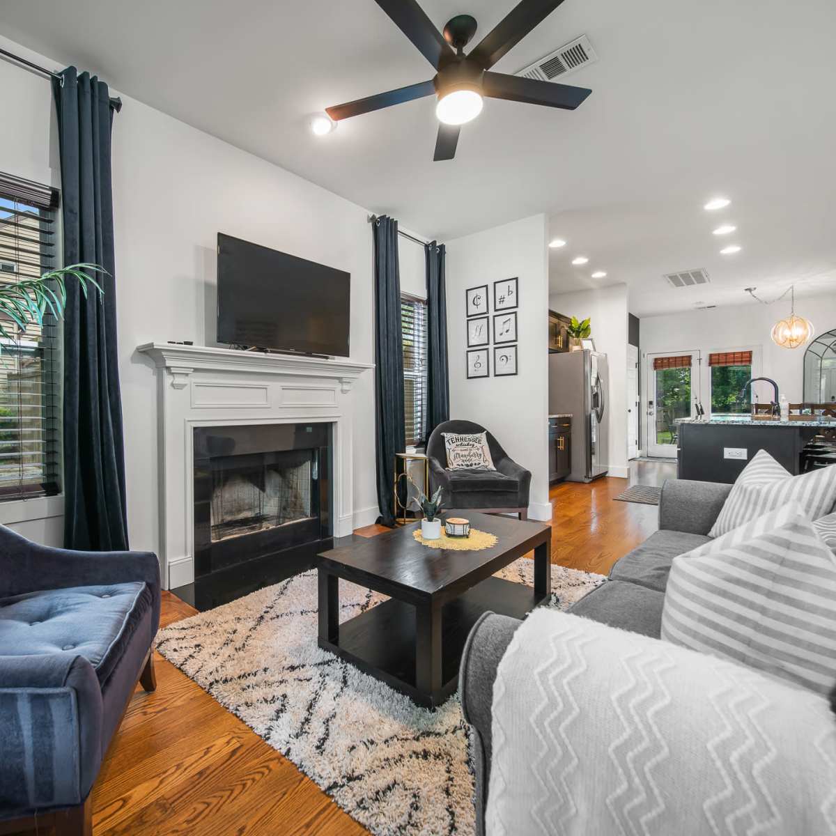 A cozy living room features a fireplace, a dark blue armchair, gray sofas, and light hardwood flooring, with large windows dressed in dark curtains.