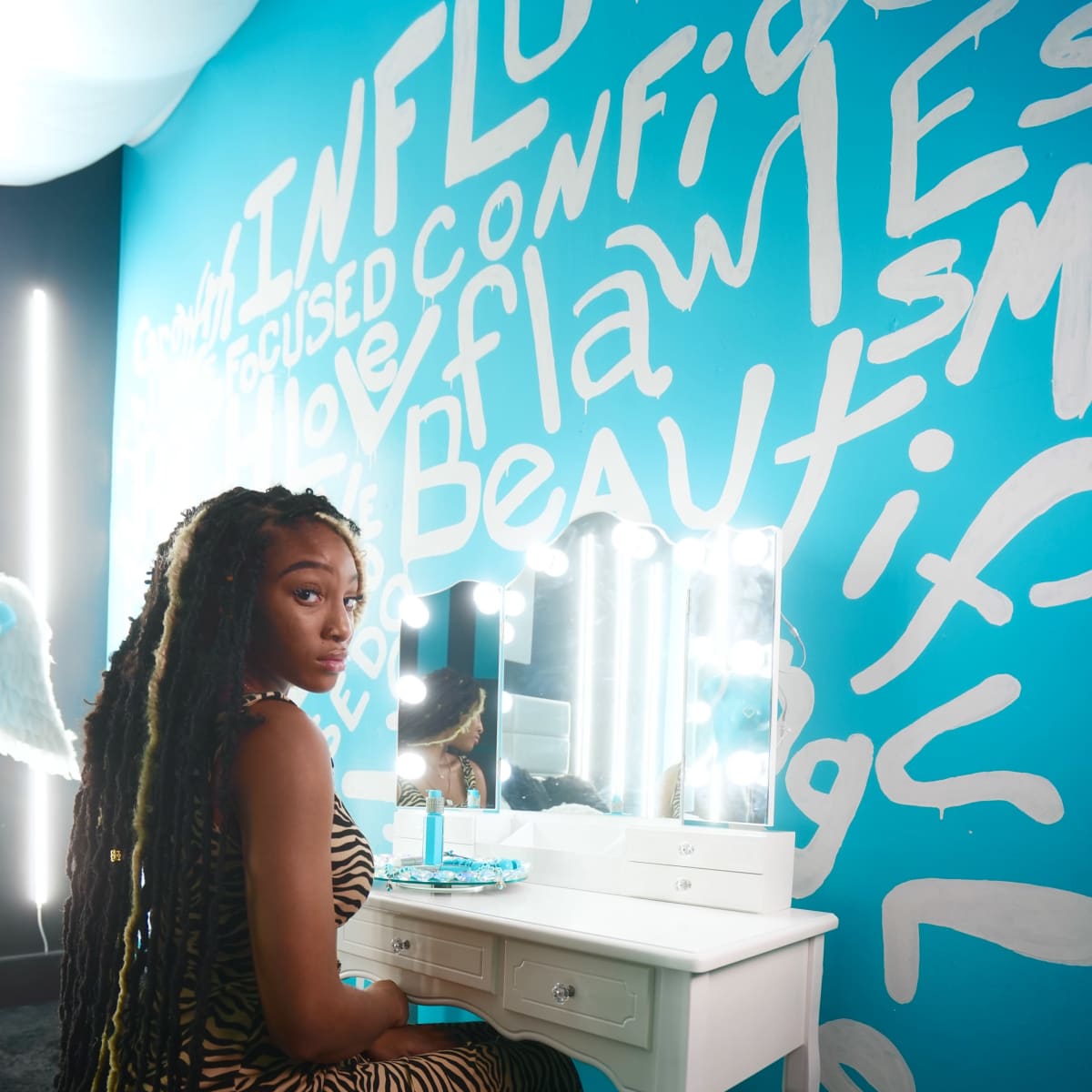 A person with long braided hair sits at a white vanity in front of a blue wall adorned with colorful text.