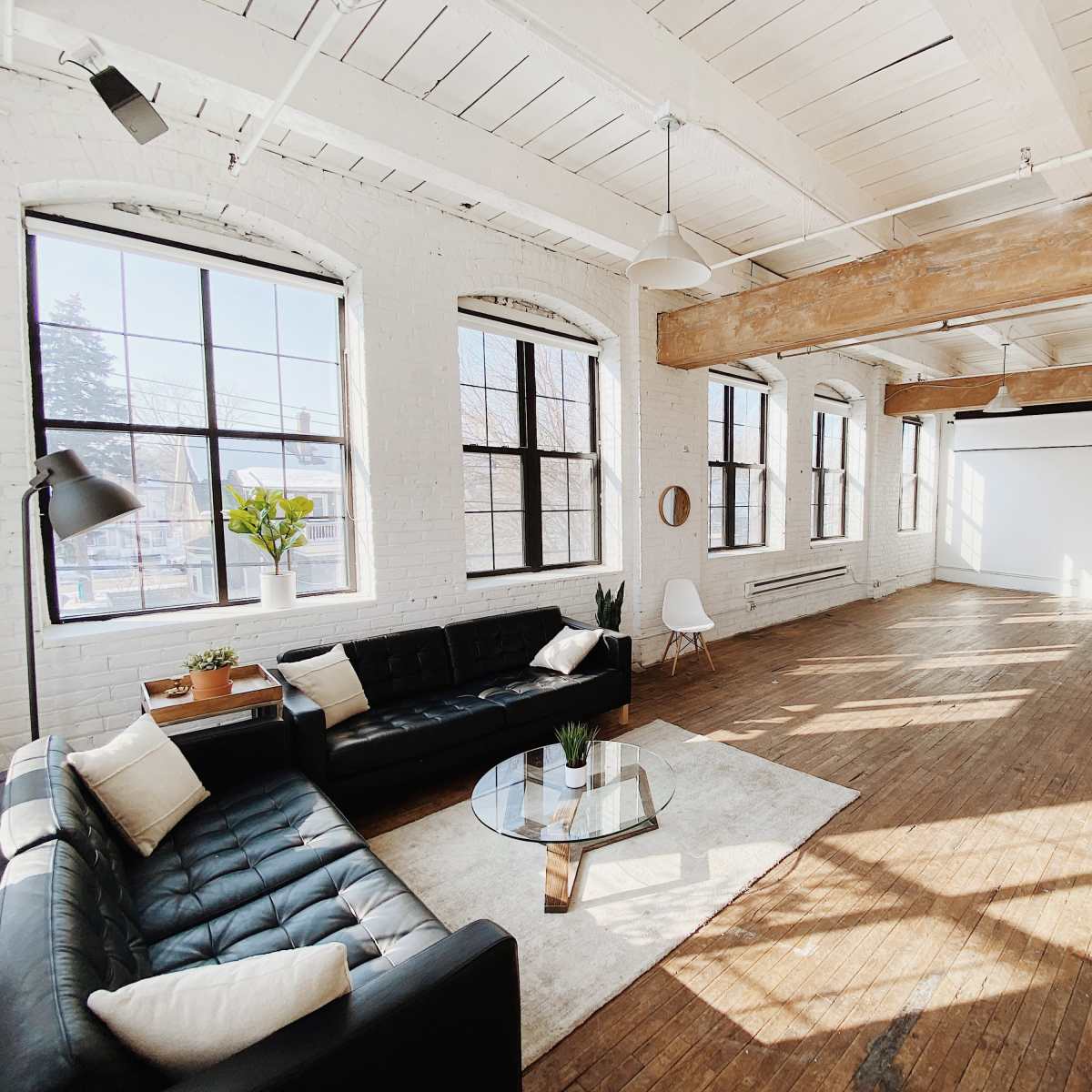 A spacious, well-lit loft with large windows, featuring a black leather sofa, a glass coffee table, and wooden flooring.
