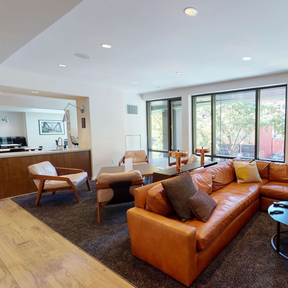 A modern living room with a brown leather sofa, a black coffee table, and large windows offering a view of trees outside.