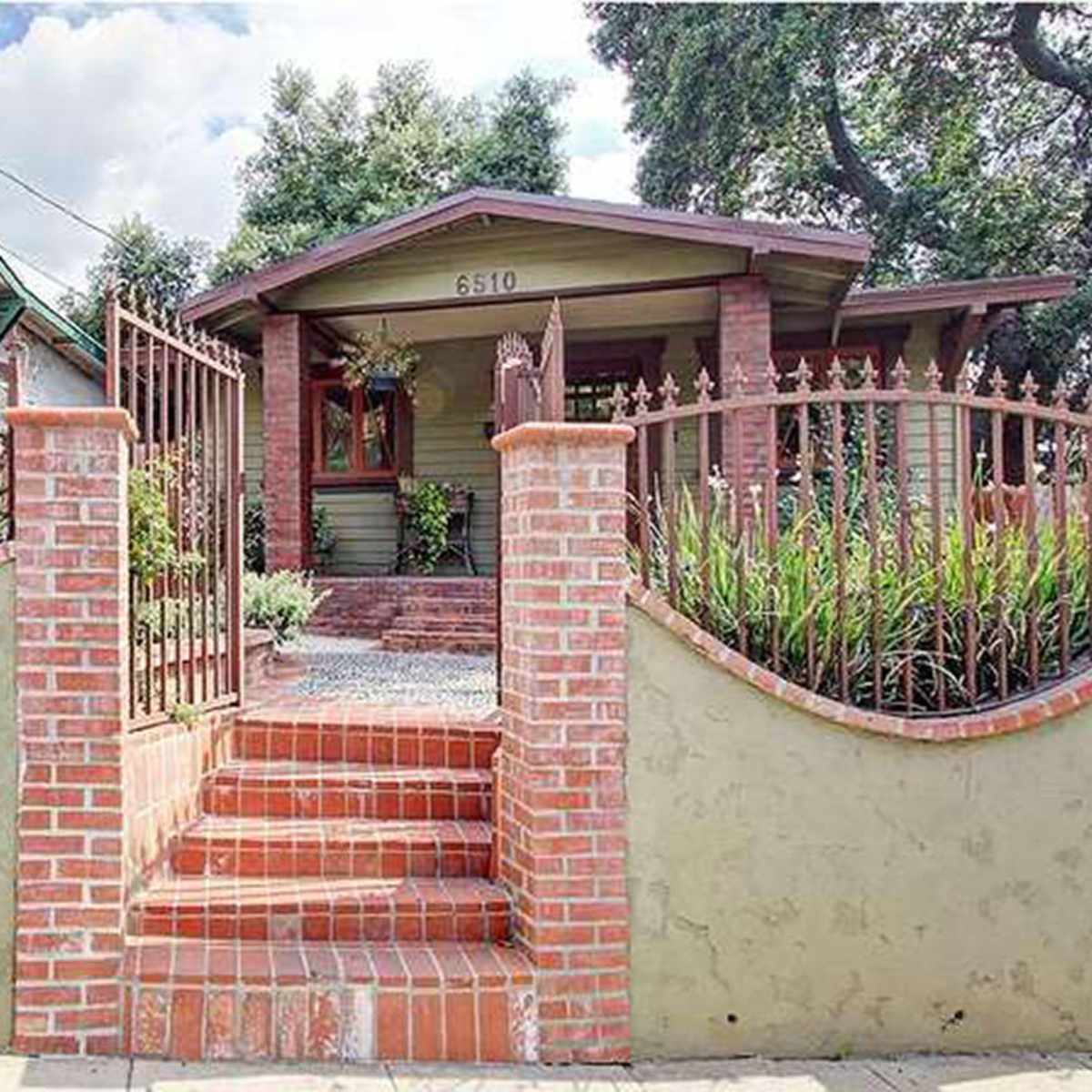 A brick-walled entrance with stairs leading to a front porch of a house surrounded by greenery.