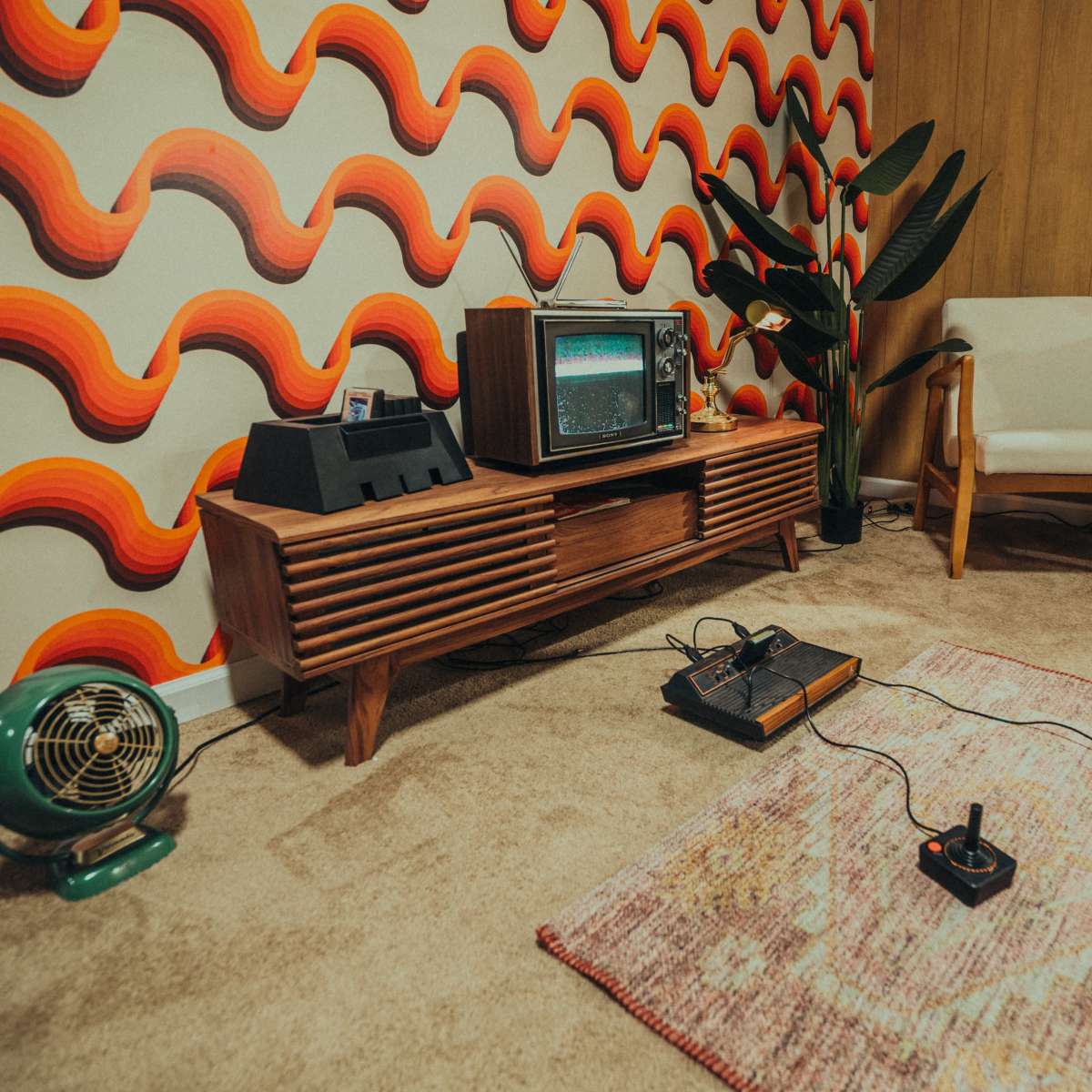 A retro living room with a vintage television, a wooden TV stand, an Atari console connected to two controllers, a green fan, and a patterned wallpaper featuring wavy orange and yellow shapes.