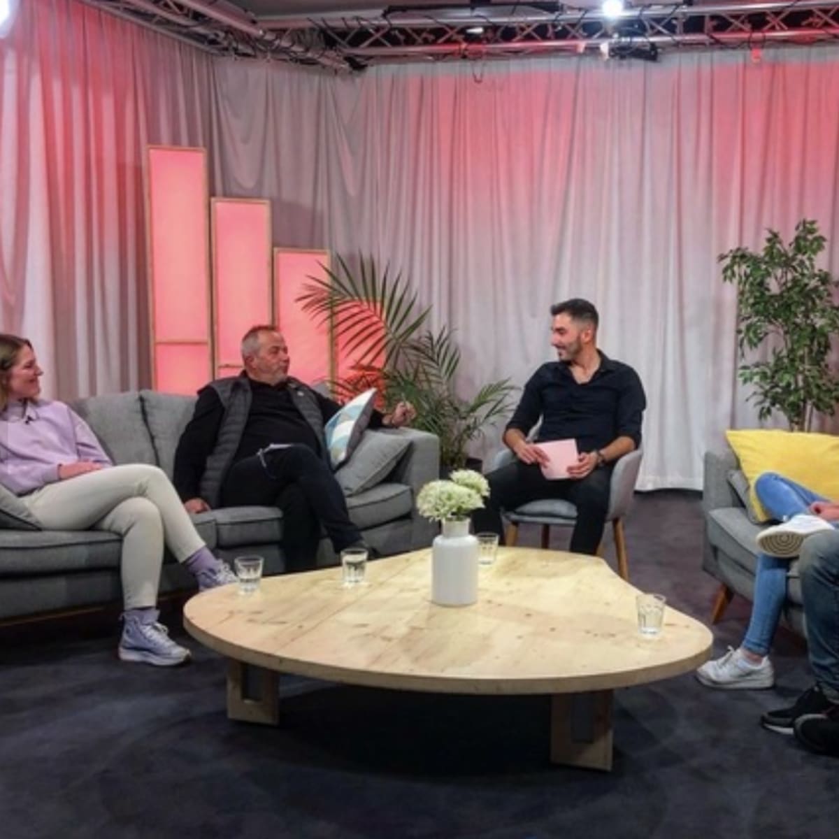 A group of six people is seated on a set with modern gray sofas and a wooden coffee table, engaged in a conversation under soft lighting and backdrop decorations.