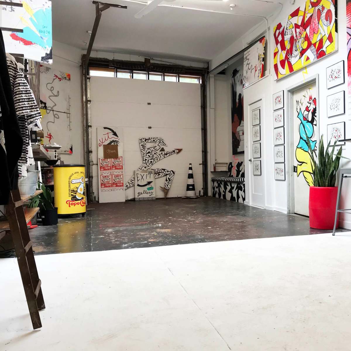 An art studio with an open space featuring colorful wall art, a large garage door, and a partially visible plant in a red pot.