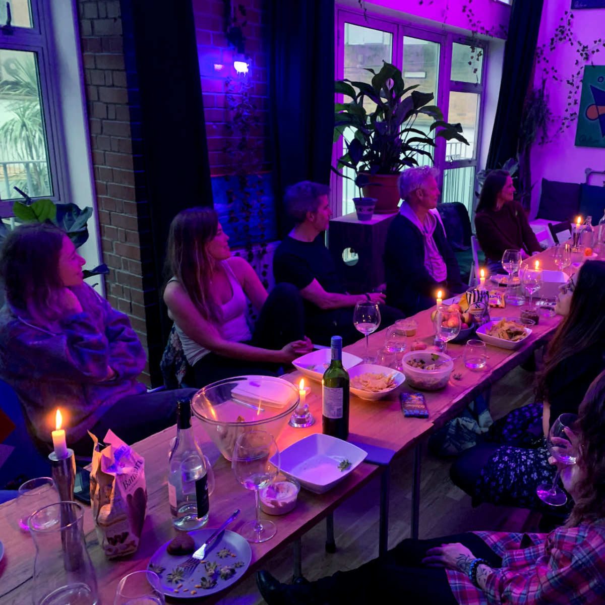 A group of people sits around a long table decorated with food and drinks in a dimly lit room filled with plants and colorful artwork.