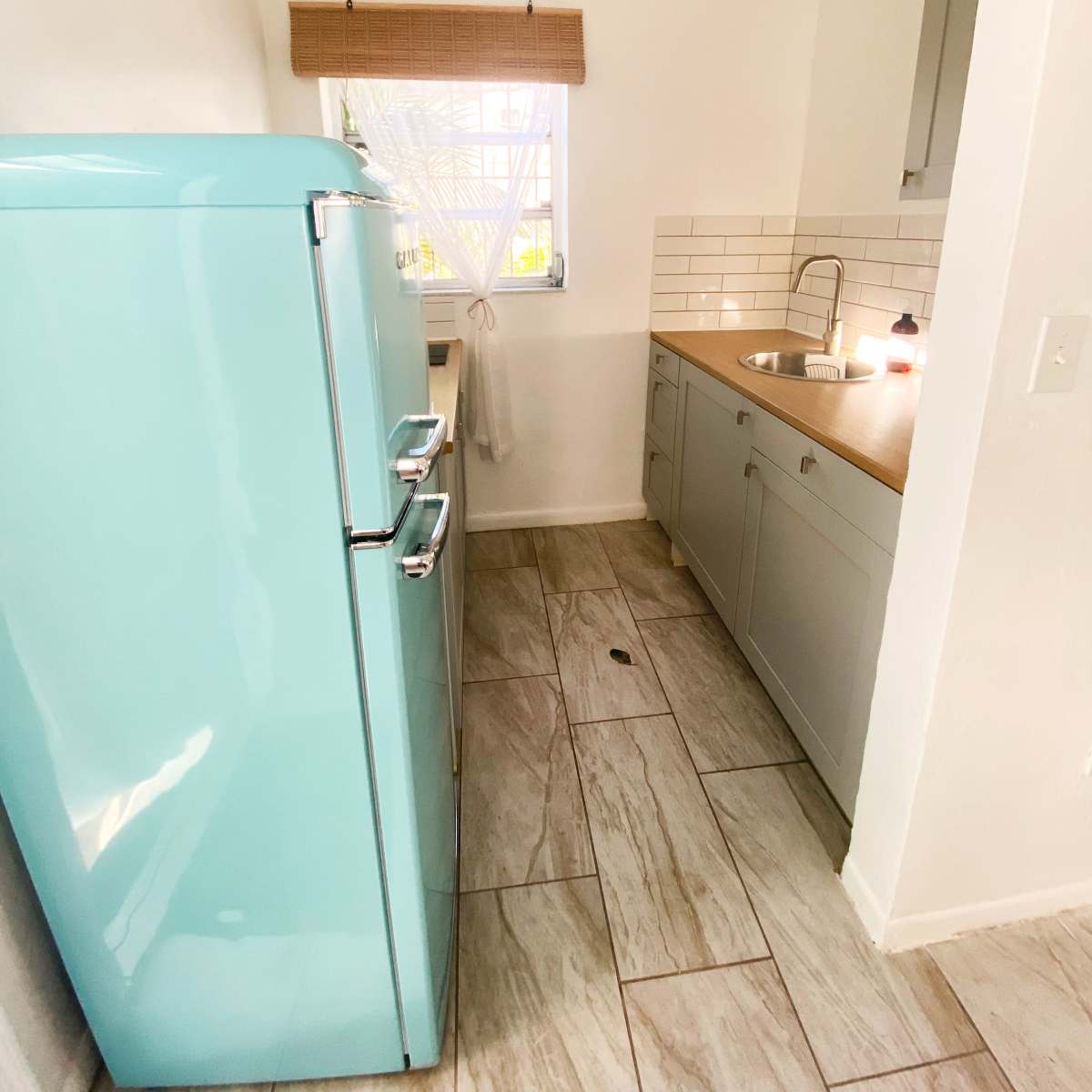 A light blue vintage refrigerator stands next to a modern kitchen sink and countertop with white tiles in a small, brightly lit kitchen space.