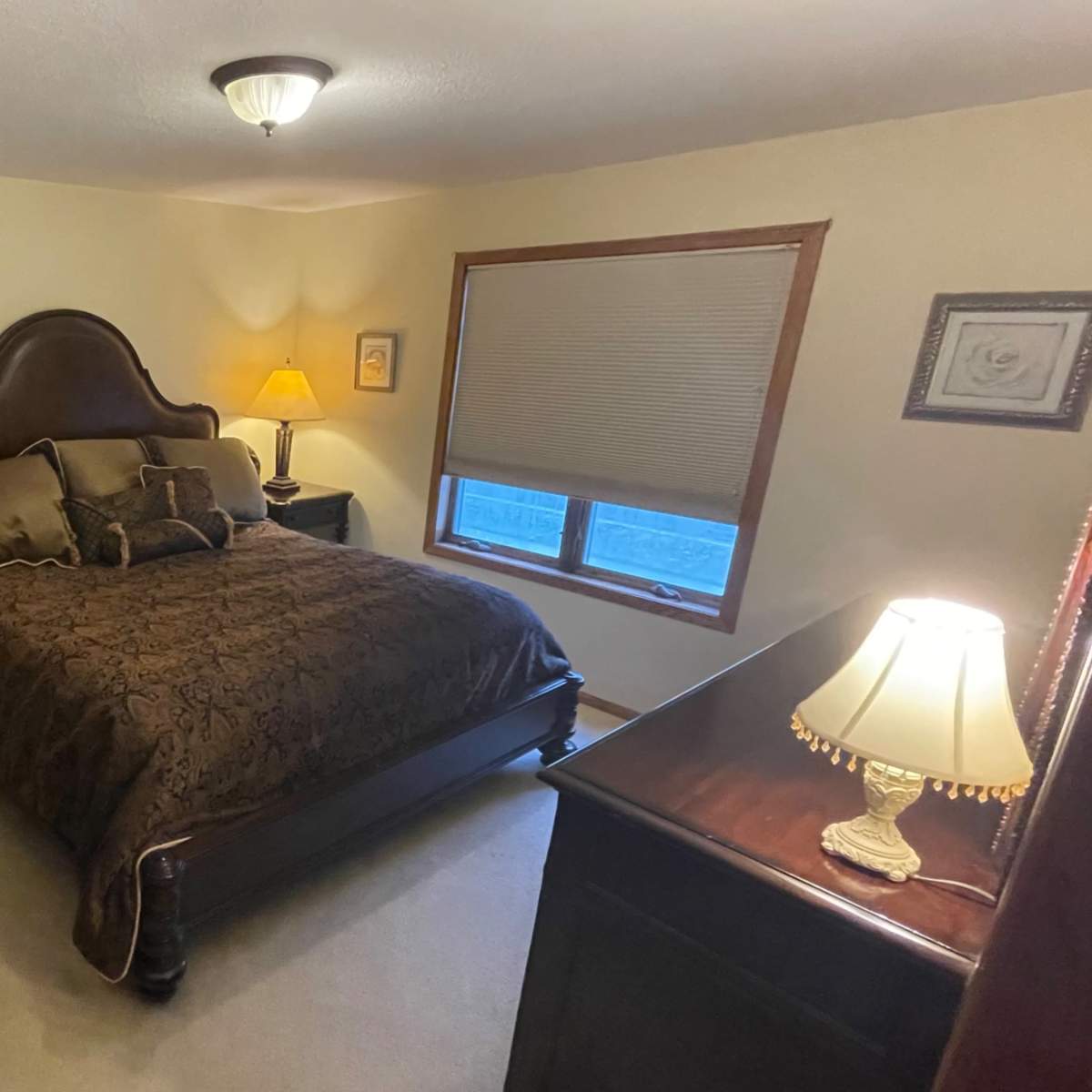 A cozy bedroom featuring a dark wooden bed with a patterned brown bedspread, a nightstand with a lamp, and a dresser against the wall.
