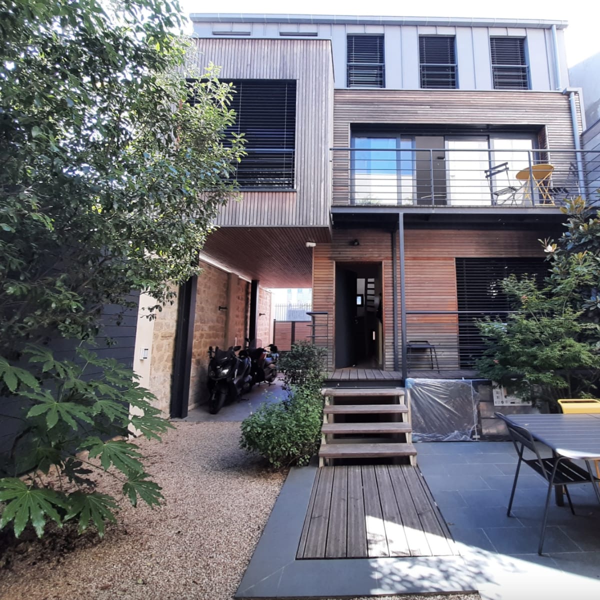A modern house with a wooden exterior, a patio area, and a garden surrounded by plants and gravel pathways.