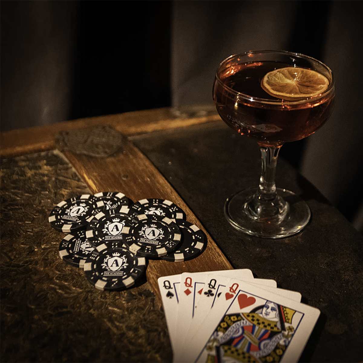 A cocktail glass with a slice of lemon sits next to a fan of playing cards and a stack of black poker chips on a wooden table.